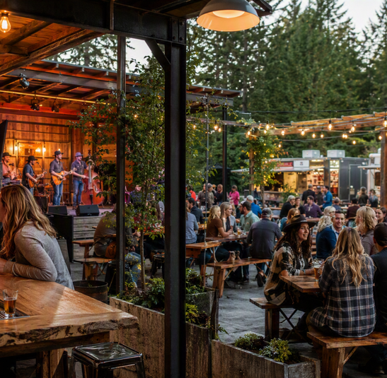 A local band playing on an outdoor stage as guests listen from picnic tables under string lights, with evergreen forest behind